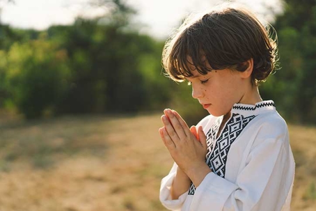 A young child wearing a traditional embroidered shirt quietly prays outdoors