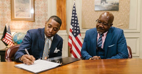Two men in suits are seated at a table, one signing a document.