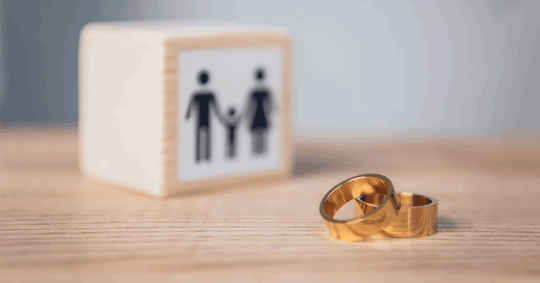 A small wooden block with a family symbol, and two gold wedding rings on a light brown surface.