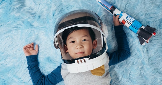 A child wearing an astronaut helmet and holding a toy rocket lies on a light blue fuzzy surface