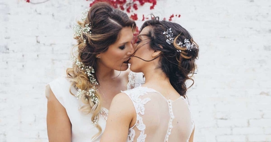 Two women in white wedding dresses are kissing.