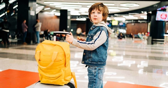 A young boy, wearing a denim jacket, is standing near a yellow backpack on a rolling suitcase in an airport.