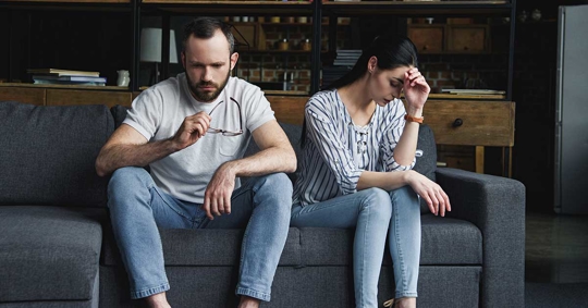 A man and a woman are sitting apart on a couch, looking distressed.