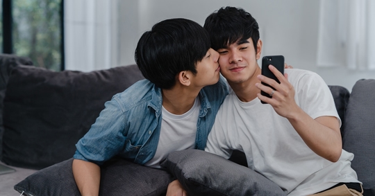 Two men are sitting on a couch, one kissing the other's cheek, and one holding a phone.
