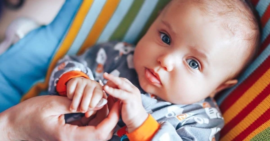 A baby, wearing a gray onesie, is lying in a blue and orange striped carrier.