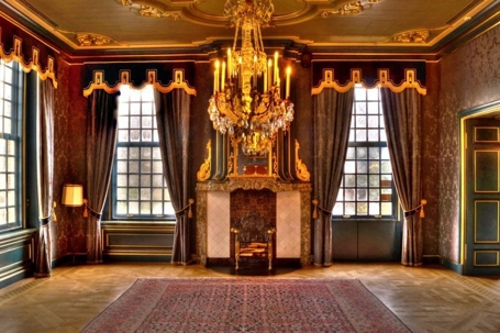 A large room with dark drapes, a fireplace, a chandelier, and a patterned rug.