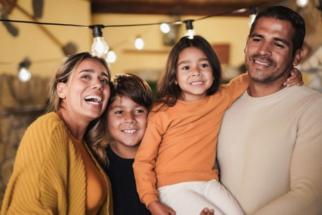 A happy family of four, a woman, man, boy, and girl, are smiling at the camera. Warm lighting.