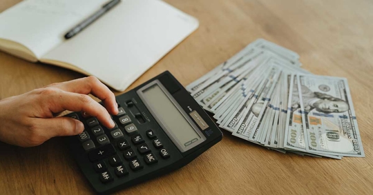 A hand on a calculator, money, and a notebook are on a wooden table.