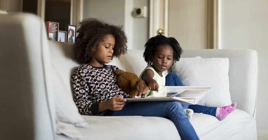 Two girls are sitting on a couch, looking at a photo album. A teddy bear is also visible.