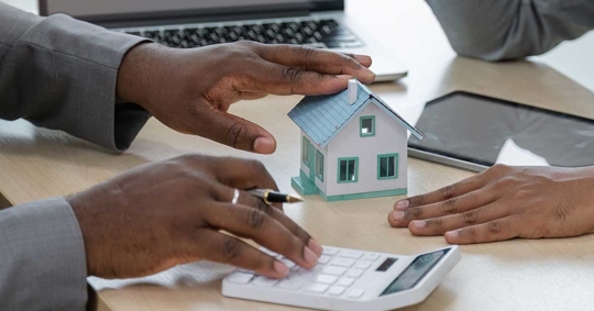 Two people are looking at a small model house, a calculator, and laptops.