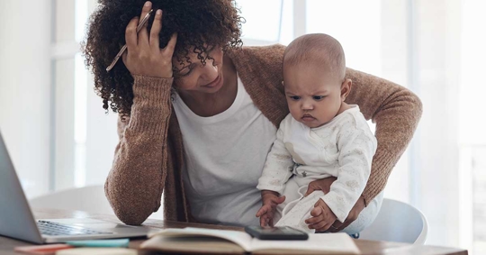 A woman looks stressed, holding a baby, and seated at a table with a laptop and papers.