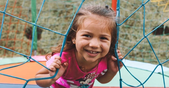 A young girl with brown hair, wearing a pink shirt, is looking at the camera through a blue netting.