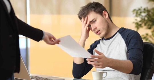 A person in a gray/navy raglan shirt is looking at a document handed to them by someone in a black suit.