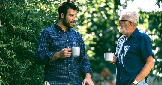 Two men, outdoors, are talking while holding mugs.