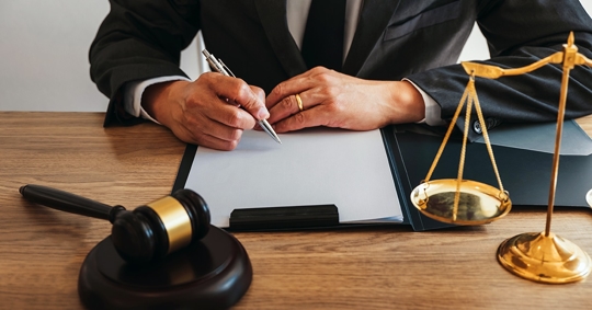 A person in a suit is writing on legal documents, with a gavel and scales of justice on a wooden table.
